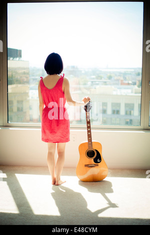 Back view of young woman standing with guitar and looking through window Stock Photo