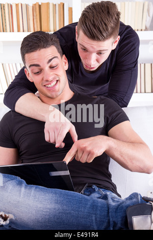 two surprised smiling friends sitting on the couch and showing how they use the tablet or laptop computer in living room atmosphere Stock Photo