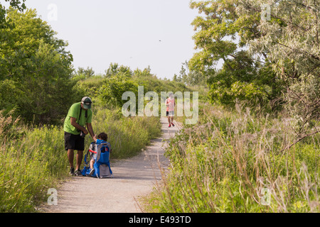 Father with toddler son in a backpack sitting on the ground on a trail as a jogger is coming by Stock Photo
