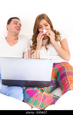 ordinary casual young couple in love watching sad movie in bed on laptop computer. Man laughing at woman with tissue while she is crying and shedding tears Stock Photo