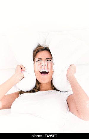 Beautiful young screaming business woman lying in bed and covering her ears with pillows. Indoor background, noise or insomnia concept Stock Photo