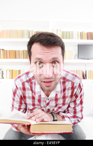 Closeup portrait of a confused shocked and happy student or professor with book in hands in living room with library behind him, nerdy, goofy, book worm Stock Photo