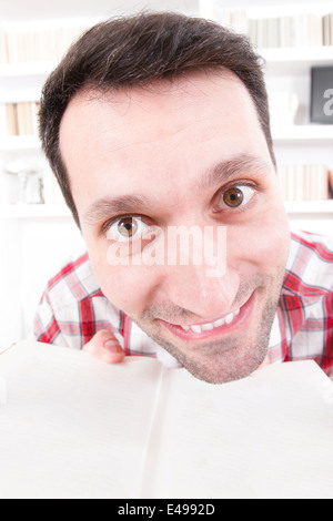 educational theme, close up of nutty confused and shocked wide eyes open professor or student with book in his hands, living room atmosphere with library background. Mad man because excessive learning or styding Stock Photo