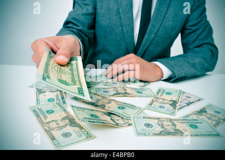 man in suit sitting in a desk full of dollar bills offering one of them to the observer Stock Photo