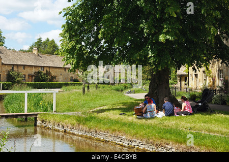 People sitting under a tree having a picnic along the river Eye in the centre of the village, Lower Slaughter, England, UK. Stock Photo