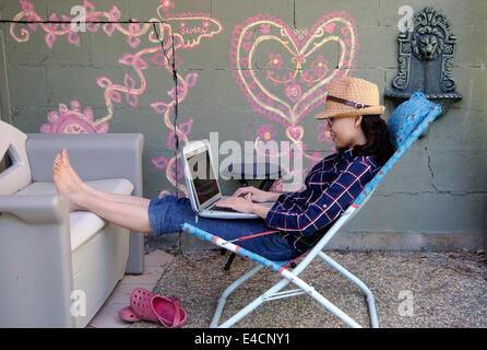 barefoot woman in straw hat  relaxes on patio in lawn chair  with laptop Stock Photo
