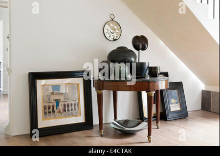 Assorted ceramics on top of marble topped round table underneath staircase with unhung pictures Stock Photo