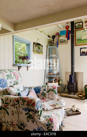 A floral patterned armchair in a cosy shepherds hut with a 'Wendy' woodburning stove by Windy Smithy Stock Photo