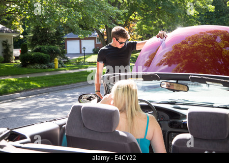 A young man looks under the hood of a red convertible car as a young woman in the car calls for assistance Stock Photo