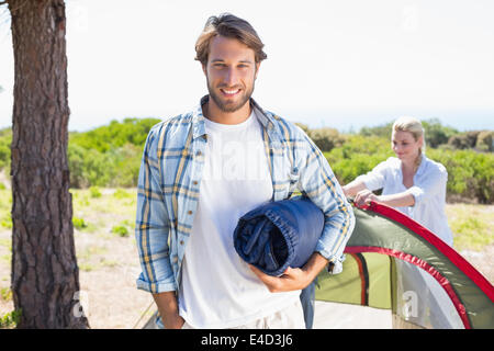 Attractive man smiling at camera while partner pitches tent Stock Photo