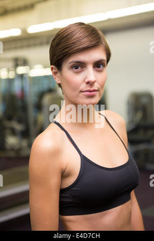 Fit brunette in black sports bra smiling at camera at the gym