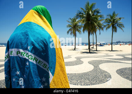 Embarrassed Brazilian wrapped in flag regalia standing in front of Copacabana Beach city skyline Rio de Janeiro Brazil Stock Photo