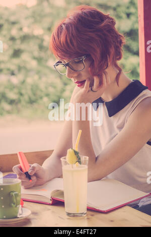 Cute hipster teenage girl daydreaming in a coffee shop, drawing hearts in her notebook, Stock Photo
