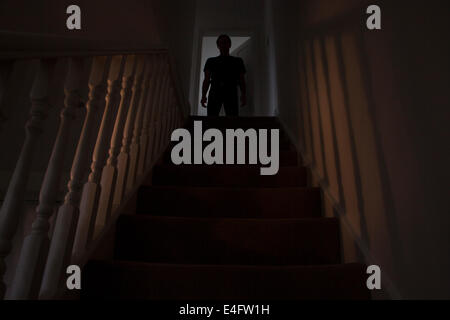 Silhouette of a man standing at the top of a stairway, shadows cast on the walls from the light below. Stock Photo