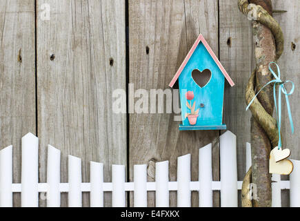 Teal blue and pink birdhouse hanging over white picket fence next to honey locust tree with hearts Stock Photo