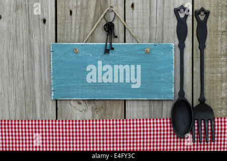 Blank antique blue sign with black iron keys hanging next to red checkered tablecloth and cast iron spoon and fork Stock Photo