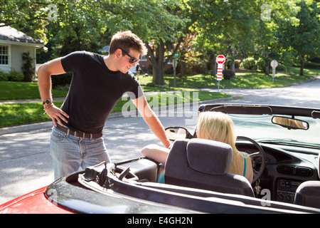 A young woman driving a red convertible stops to talk to a young man on the road in a residential neighborhood. Stock Photo