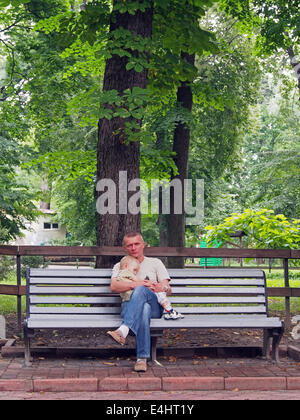 man sitting on a bench under a tree and holding on his lap sleeping son Stock Photo