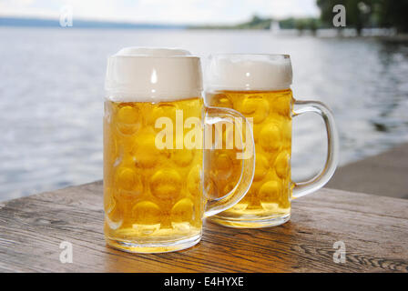 two beer mugs with fresh beer and foam on a wooden table in a beer garden on lake, close up Stock Photo