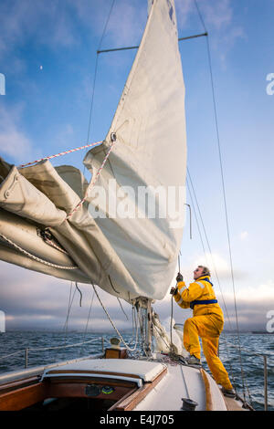 Sailor in yellow foul weather gear pulling on the halyard to raise the mainsail as he heads into cloudy weather in his sailboat Stock Photo