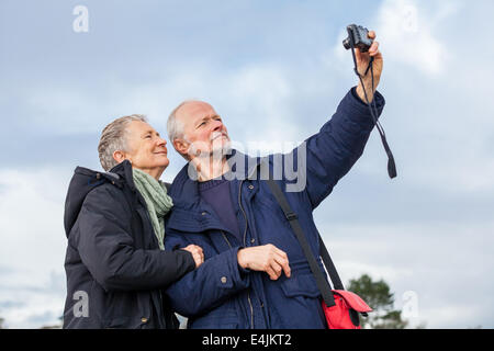 Elderly couple taking a self portrait on a compact digital camera posing in the open air and sunshine with their heads close tog Stock Photo