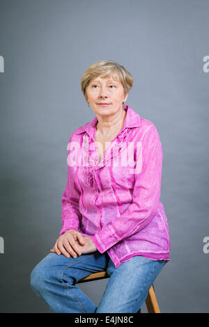 Beautiful blond trendy senior woman posing in a pink blouse and jeans sitting on the floor clasping her knee and smiling at the Stock Photo