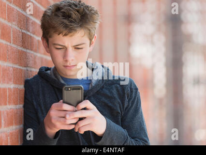 Teenage boy (16-17) leaning against brick wall texting Stock Photo