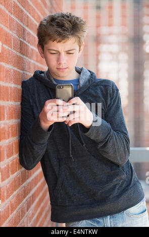 Teenage boy (16-17) leaning against brick wall texting Stock Photo