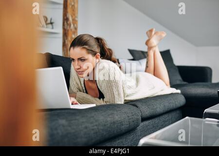Image of young woman busy using a laptop at home. Caucasian female model lying on couch working on laptop computer. Stock Photo