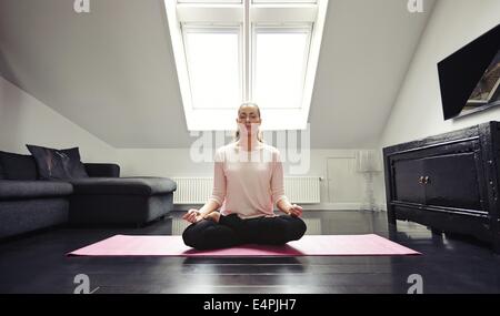 Portrait of young woman meditating in lotus position at home. Caucasian female model sitting with legs crossed on living room fl Stock Photo