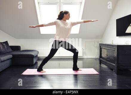 Portrait of a young woman doing yoga on an exercising mat in her living room. Caucasian female model in warrior pose.