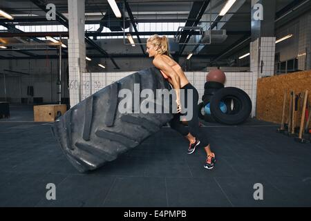 Fit female athlete flipping a huge tire. Muscular young woman doing crossfit exercise at gym. Stock Photo