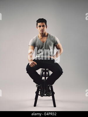 Portrait of smart young man sitting on stool. Attractive male model posing over grey background. Stock Photo