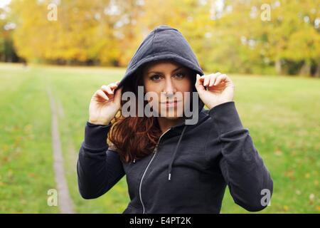 Portrait of beautiful young woman in sportswear looking at camera. Confident female runner in hoodie standing outdoors in park. Stock Photo