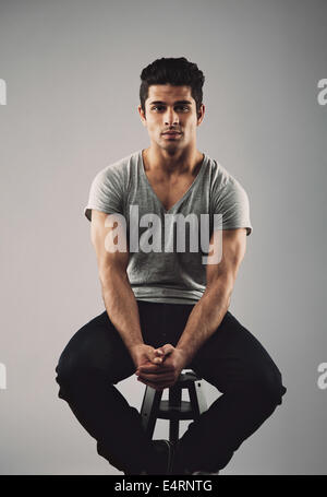 Portrait of muscular young male model sitting on stool. Hispanic young man over grey background. Stock Photo