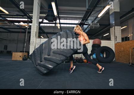 Fit female athlete working out with a huge tire, turning and flipping in the gym. Crossfit woman exercising with big tire. Stock Photo