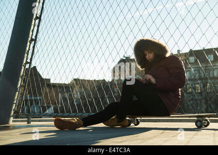 Teenage girl sitting on skateboard next to chain link fence near comercial dock, wearing winter coat, using smart phone, Germany Stock Photo