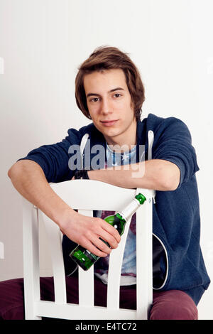 Portrait of teenage boy sitting on chair holding bottle of beer, smiling and looking at camera, studio shot on white background Stock Photo