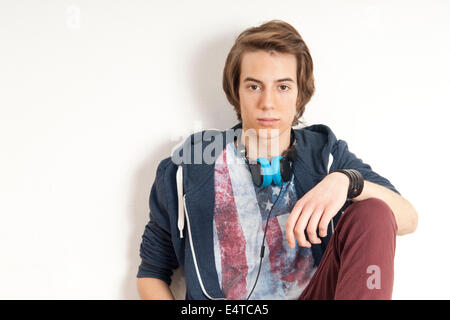 Portrait of teenage boy leaning against wall, with headphones around neck, looking at camera, studio shot on white background Stock Photo