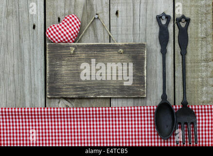 Blank rustic sign with red checkered (gingham) heart and tablecloth hanging next to cast iron spoon and fork Stock Photo