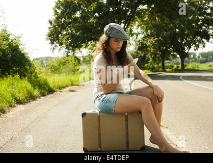 Teenage girl sitting on suitcase on the side of the road, looking at cell phone in summer, Germany Stock Photo