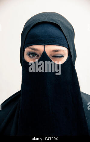 Portrait of young woman in black, muslim hijab and muslim dress, winking at camera, eyes with makeup, on white background Stock Photo