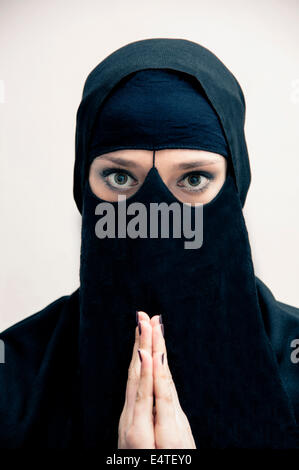 Portrait of young woman in black, muslim hijab and muslim dress, with hands praying, looking at camera, on white background Stock Photo