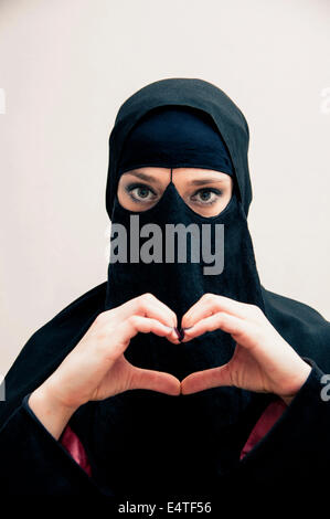 Portrait of young woman in black, muslim hijab and muslim dress, making heart shape with hands, on white background Stock Photo