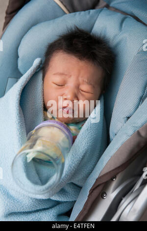Close-up of swaddled two week old, newborn Asian baby girl, lying in car seat, disgusted with her milk bottle Stock Photo