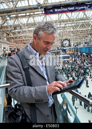 iPad Railway station Mature businessman at London Waterloo railway station using his iPad air tablet computer 'on the move' with commuters behind Stock Photo