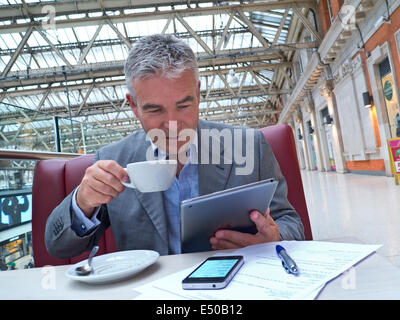 Mature businessman seated at cafe table on railway concourse looking at his tablet computer with iPhone and notes in foreground Stock Photo