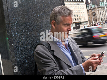 Mature businessman using an iPhone in London Southbank with blurred traditional London black cab passing in background Stock Photo