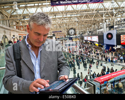 Mature businessman on London Waterloo railway concourse using his iPad air tablet computer 'on the move' with commuters behind Stock Photo
