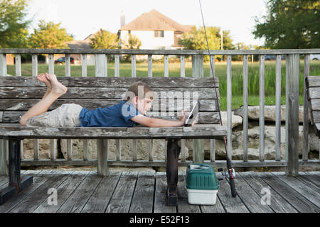 A young boy outdoors lying on a bench using a digital tablet. fishing equipment. Stock Photo